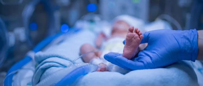 Recém-nascido prematuro em incubadora de UTI neonatal, com o pezinho segurado pela mão enluvada de um profissional de saúde — ilustração sobre prematuridade e a importância de prevenir o parto prematuro